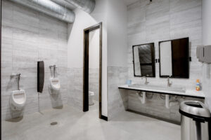 View of the men's restroom inside of 105 Nursery Lane with grey tiled floors and walls, a sink, wooden mirrors, and urinals