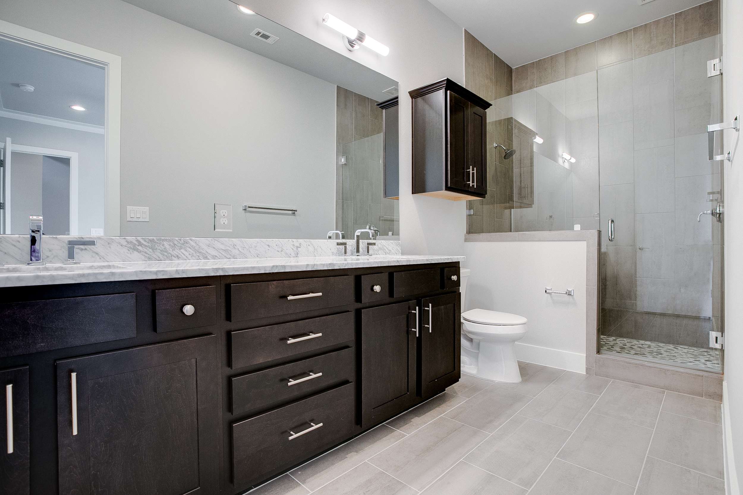 Interior view of the bathroom with dark cabinets, double sinks, a toilet, and a tiled shower with a glass door
