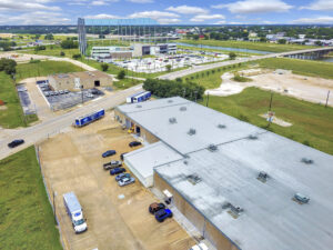 Aerial view of a tan building with a grey roof and a lot of trailers and cars with views of Top Golf in the background
