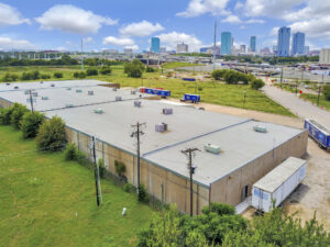 Aerial view of the backside of a tan building with a grey roof with power lines and a view of downtown Fort Worth