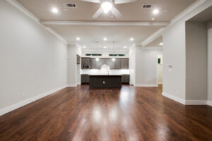 Interior view of the empty living room with dark wood floors, white crown molding and a view into the kitchen