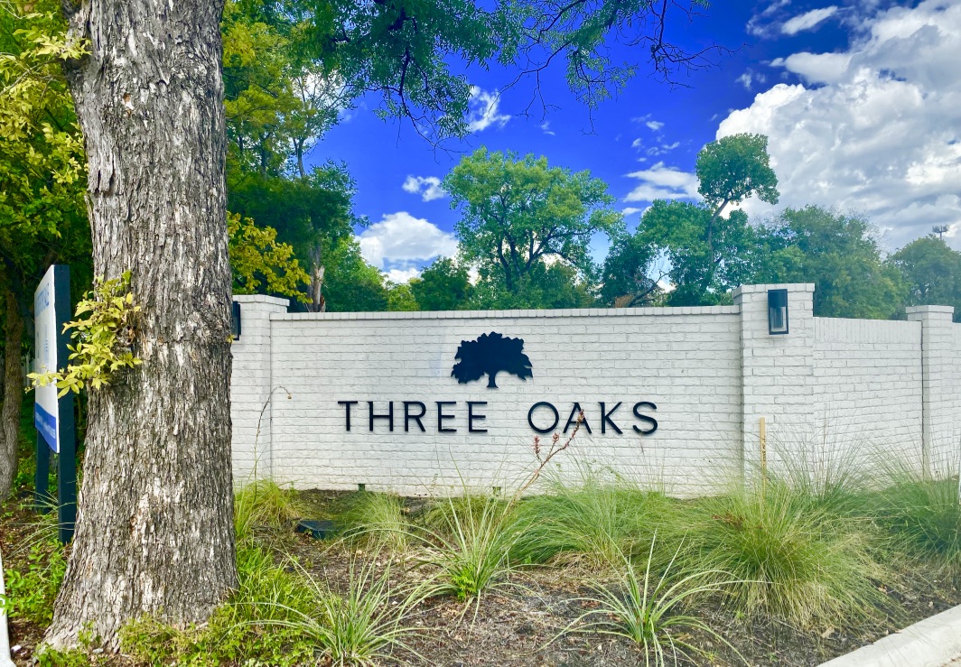 Up-close view of black lettering saying "Three Oaks" on a white brick fence with a trunk of a brown tree on the left