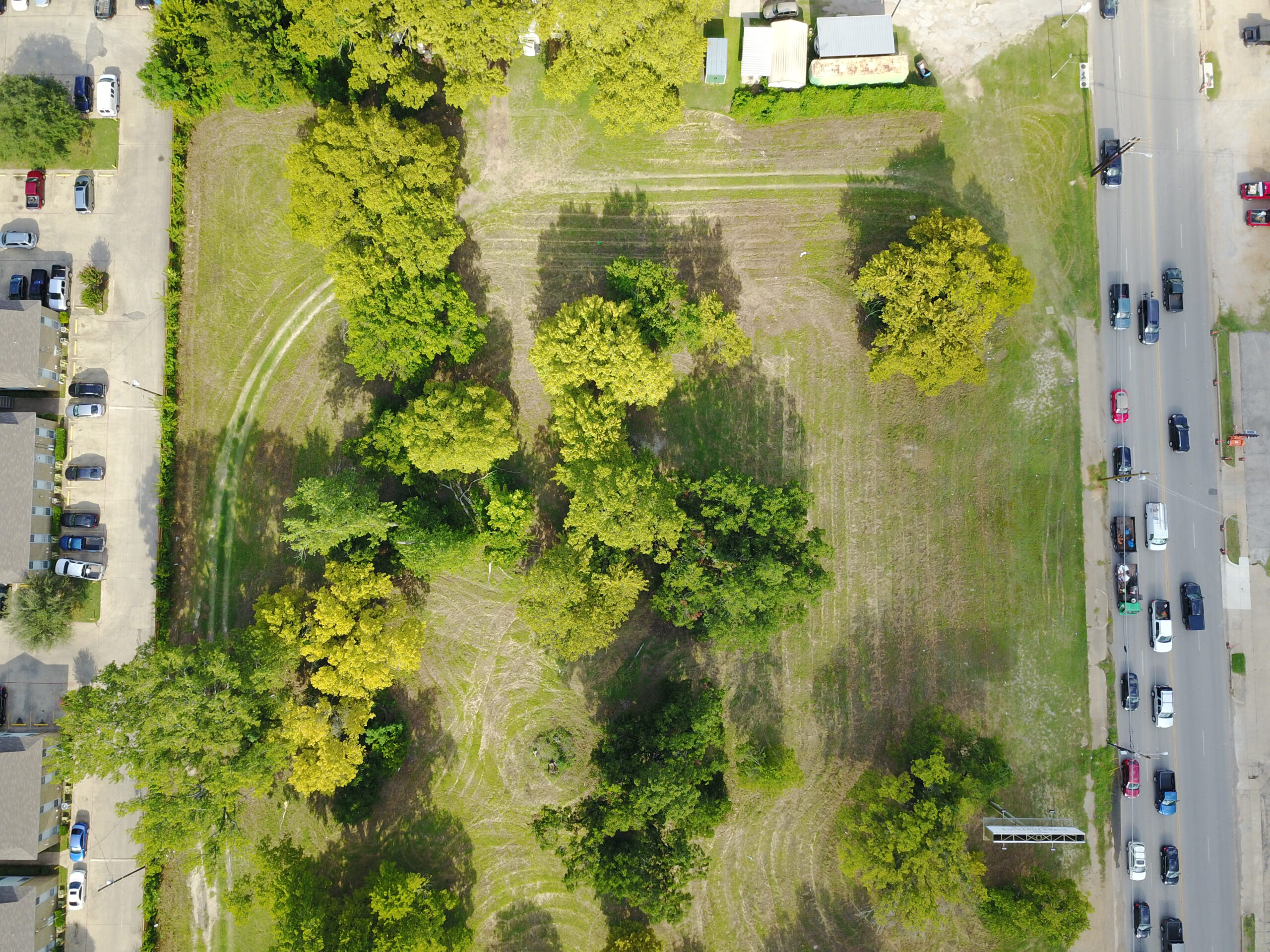 Birds-eye view of a grassy plot of land with trees adjacent to a parking lot on one side and a street on the other