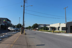 Ground-level view of a street with a stop sign, a grey building on the right, a parking lot on the left, and an electric line